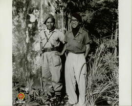 Dua orang gerilyawan bersiaga berdiri di samping tembok di antara pepohonan.